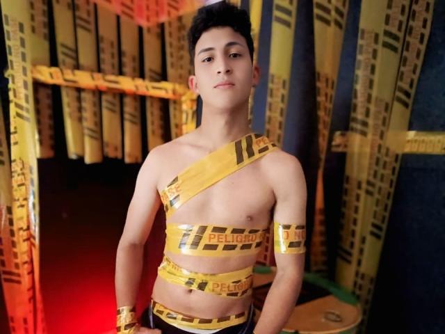 AxelSavage, avec un corps mince et musclé d'un garçon asiatique, portant du ruban jaune "PETROCORD" enroulé autour de sa poitrine, de son bras et de sa taille, se tient dans une pièce sombre éclairée en rouge..