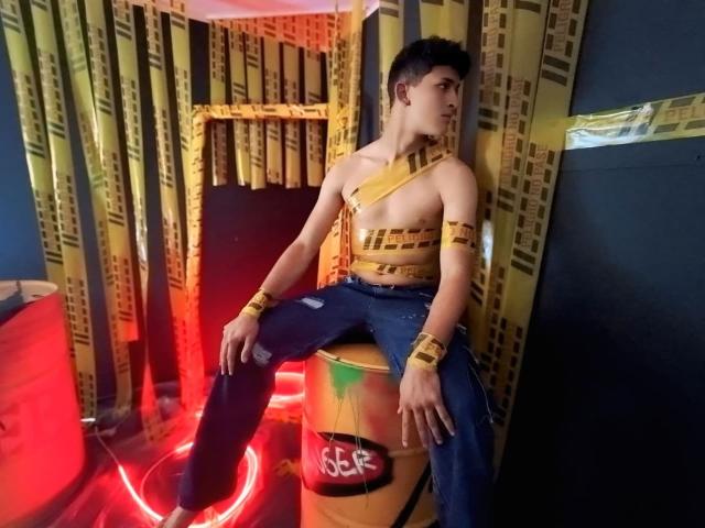 AxelSavage, avec du ruban jaune enroulé autour de sa poitrine nue et de ses bras, assis sur un baril jaune, portant un jean bleu, dans une pièce faiblement éclairée avec des lumières rouges et jaunes..