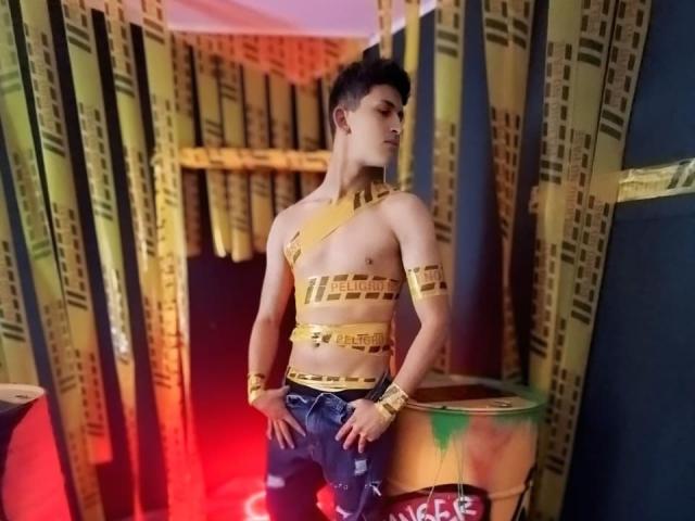 AxelSavage, senza camicia con del nastro dorato che copre il petto e le braccia, indossa jeans blu strappati, è in piedi in una stanza poco illuminata, ha i capelli corti e scuri, le mani sui fianchi e guarda di lato..