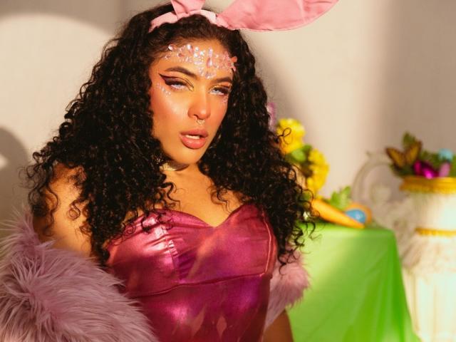 VioletHotX with curly black hair, pink bunny ears, shiny purple dress, pink glitter on face, fluffy purple arm, looking sexy, bright light, colorful background, pink lips, seductive expression.