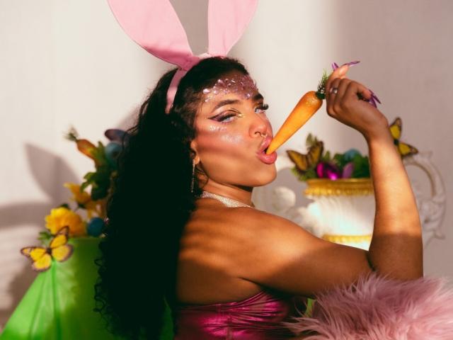 VioletHotX with long black curly hair, pink bunny ears, eating a carrot, wearing a pink strapless top, glitter on face, holding a basket of colorful eggs, butterflies.