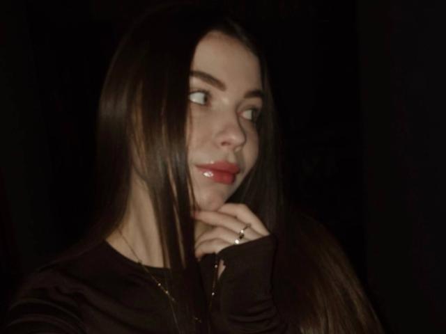 ChloeRose69 with long brown hair, glossy red lips, and a ring on her finger, looking to the side with a slight smile, dark background, wearing a black shirt.