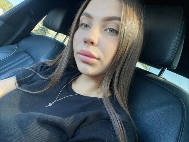 ChloeRose69 with long brown hair, green eyes, and pink lips, wearing a black shirt, sitting in a car, looking at the camera, wearing a silver necklace.