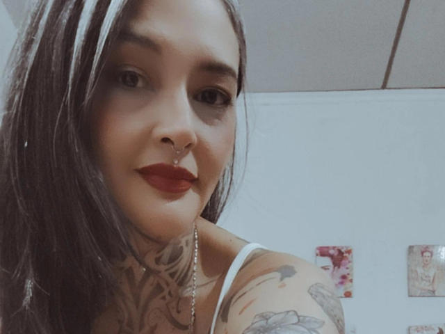 Inhara, cheveux foncés, anneau au nez, rouge à lèvres rouge, tatouages sur le cou et le bras, haut blanc, souriante, carreaux de plafond en arrière-plan, regardant la caméra, selfie légèrement incliné..