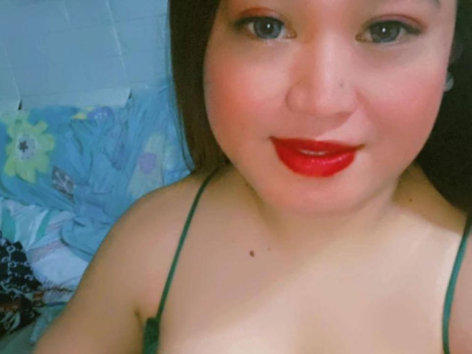 TSDelylicious69 has a chubby face, red lipstick, green spaghetti strap top, small moles on her chest, and is smiling at the camera with brown eyes.