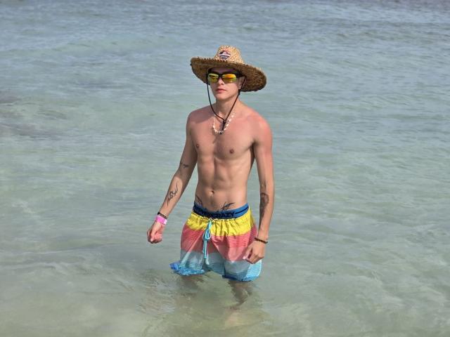 Archangel69, shirtless with a straw hat, yellow and pink swim trunks, yellow sunglasses, standing in clear water, has tattoos on both arms, and a pink wristband.