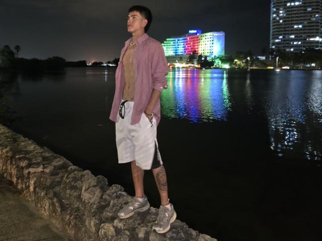 Archangel69 stands on a stone wall, wearing a pink shirt, beige tee, white shorts, and gray sneakers, with a tattoo on his right calf, at night by a reflective water.