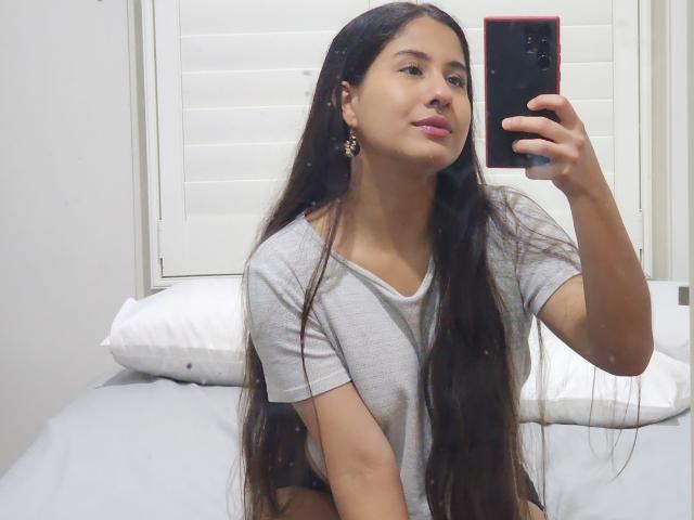 Aurora Monroe mit langen, schwarzen Haaren, die ein graues T-Shirt trägt, macht ein Selfie vor einem Spiegel mit einem roten Telefon und zeigt dabei ihren Nasenring und ihre Ohrringe, während sie auf einem Bett sitzt..