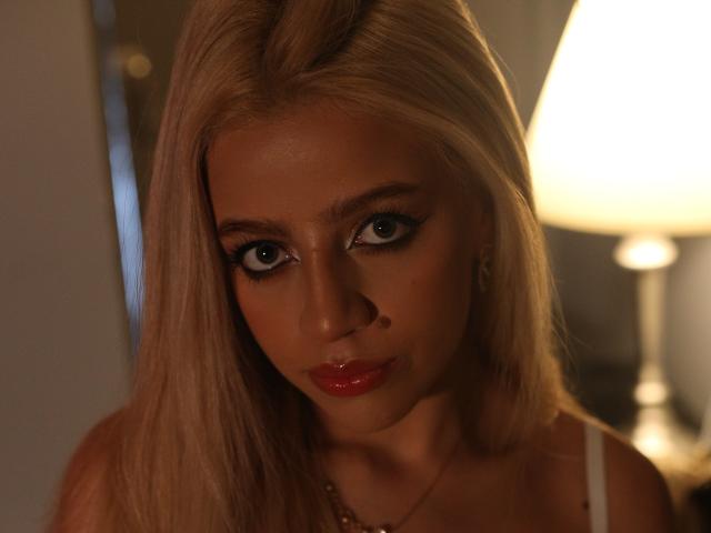 ZhaaviaMoon with shiny blonde hair, blue eyes, and glossy red lips, staring intensely at the camera in a dimly lit room, wearing a white top.
