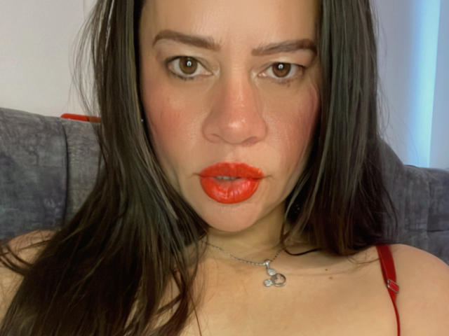 CharllotI heeft rode lippenstift, bruine ogen en steil bruin haar, draagt een rode band over haar schouder en een zilveren ketting met een klein hangertje, en ze heeft een serieuze blik..