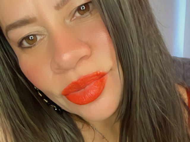 CharllotI has red lipstick, brown eyes, and straight dark brown hair, with a slightly flushed face and a subtle smile. Her tits are not visible in this shot.