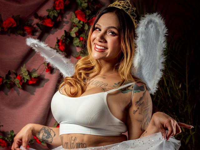 AzhunaStar with blonde ombre hair, white wings, and gold tiara, smiling, showing big tits in a white crop top, tattoos on arms and chest, red roses in background.