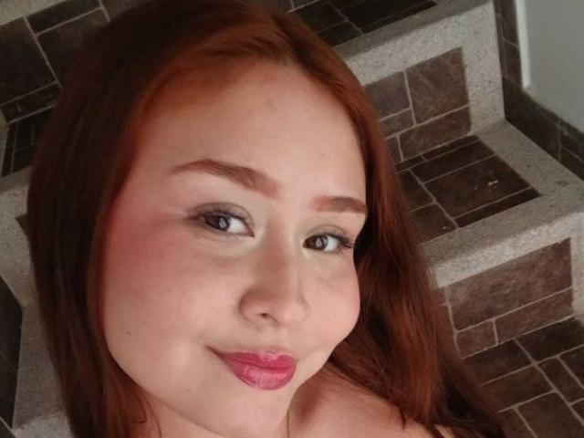 BellaMiaI has red hair, light skin, and pink lipstick with a slight smile, looking at the camera. Her tits are not visible. She's sitting on tiled stairs.