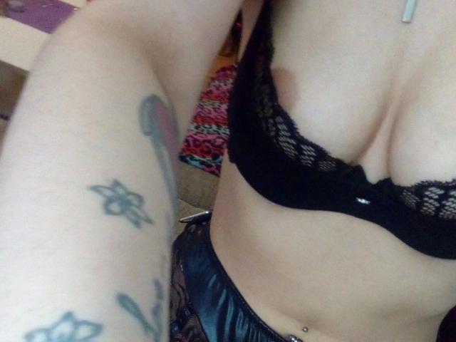 GermanVixen-hot's boobs in a black lace bra, showing a tattooed arm with a flower and a sword. She's got a belly button piercing and's wearing black panties.