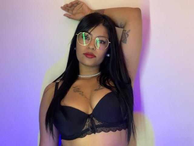 DannaHarmonyy with black bra, lace trim, and pearl necklace, wearing reflective glasses, arm raised, showing tattoos, and red lipstick, standing against a plain wall.