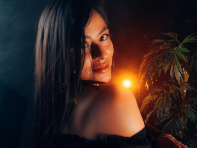 ZaiiraFox met donker, nat haar, kijkt over haar schouder, glimlachend, oranje licht op de achtergrond, blote schouders, zwarte top met een open schouder, plant aan de rechterkant, sexy, warme gloed..