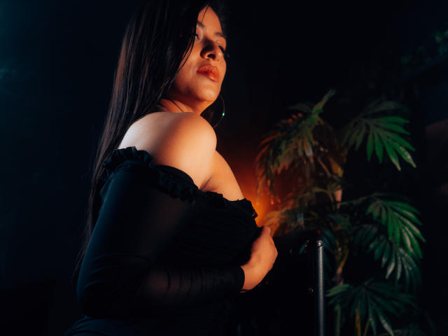 ZaiiraFox, con cabello oscuro, blusa negra con un hombro descubierto, sosteniendo sus grandes senos, mirando por encima del hombro, luz suave, planta verde, fondo oscuro, sensual, segura de sí misma, sexy, luz de la tarde..