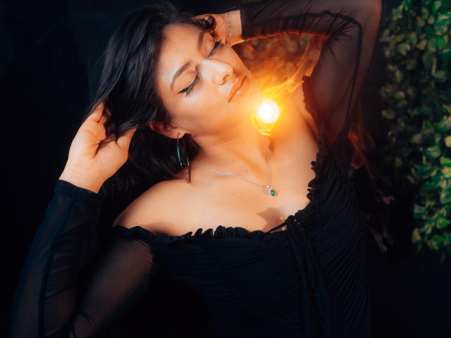 ZaiiraFox met gesloten ogen, een zwarte jurk met open schouder, en een lamp die haar gezicht verlicht, haar borsten zijn licht zichtbaar, haar hand in haar haar, groene oorbellen, een ketting, en een gloeiend licht..