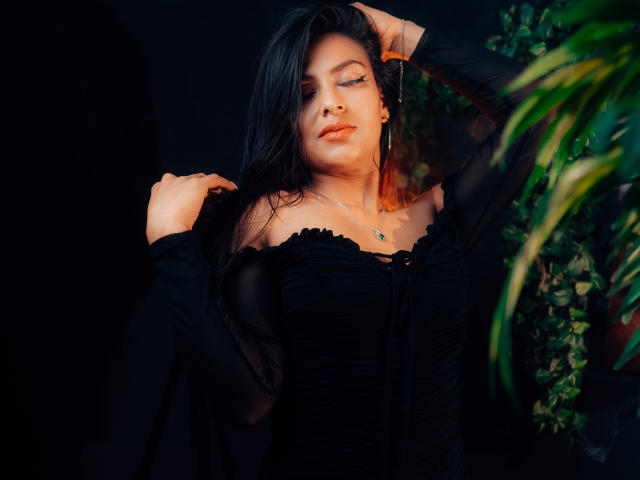 ZaiiraFox met donker haar, gekleed in een zwarte jurk met een open schouder, die haar haar aanraakt met één hand, met gesloten ogen, een zachte glimlach, een ketting, een groene plant in de rechter voorgrond..