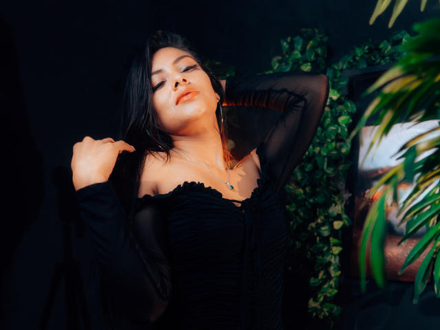 ZaiiraFox met donker haar, die een zwarte top draagt met een open schouder, handen in haar haar, ogen gesloten, een ketting, zacht licht, een groene plant en een gedeeltelijk zichtbaar poster op de achtergrond..