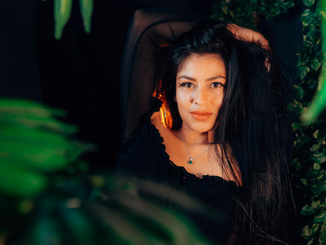 ZaiiraFox, met lang zwart haar, draagt een zwarte top met een open schouder en heeft haar hand in haar haar. Ze kijkt met een lichte frons naar de camera. Op de voorgrond staan groene planten..