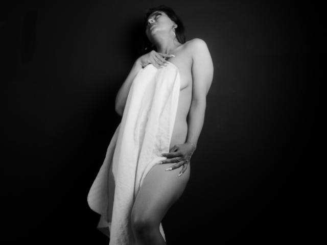 ZaiiraFox is naakt, met een witte handdoek die haar borsten en vulva bedekt. Ze houdt de handdoek vast met één hand, haar andere hand rust op haar billen, en kijkt omhoog. Zwart-wit foto..