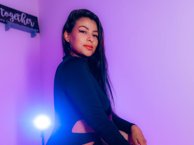ZaiiraFox met lang zwart haar, gekleed in een zwarte top met lange mouwen, zittend, kamer verlicht met paars licht, lichte glimlach, blauw licht op de achtergrond, schattig, sexy, zelfverzekerd, kijkt naar de zijkant..
