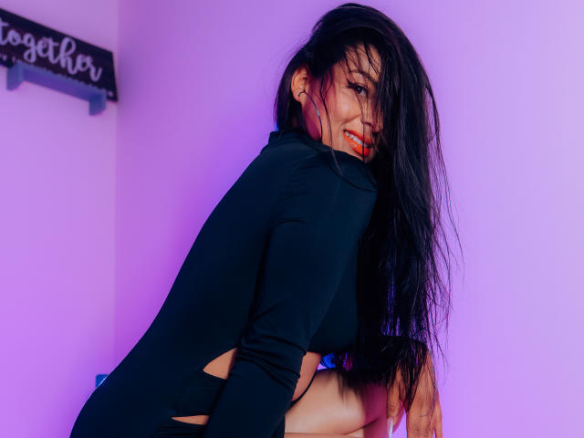 ZaiiraFox met lang zwart haar, een zwarte top met lange mouwen en uitgesneden zijkanten, knielend, lachend en over de schouder kijkend, paars en blauw licht, nat haar, rode lipstick, sexy..