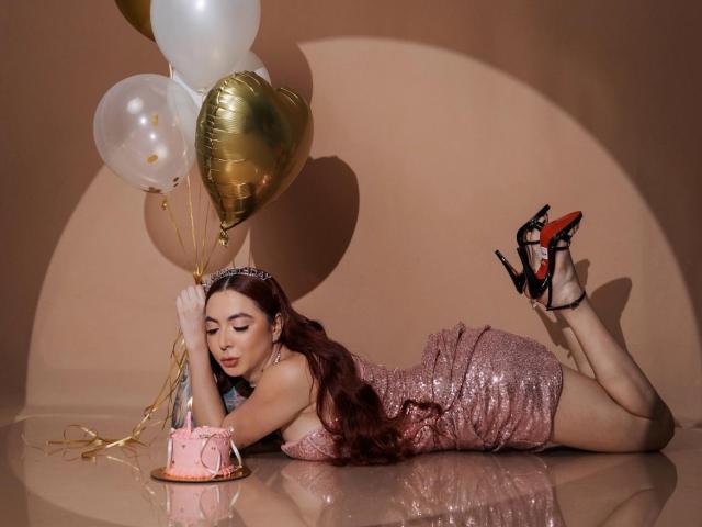 Victoria Gusman, con su largo cabello rojo, vestido rosa brillante y tacones negros, está acostada boca abajo, sosteniendo globos dorados y blancos, junto a un pastel rosa, mostrando su trasero..