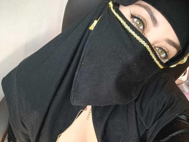 ZahraJalid with green eyes, black headscarf, and black mask, showing only her eyes and a bit of cleavage with a gold chain. Boobs partially visible.