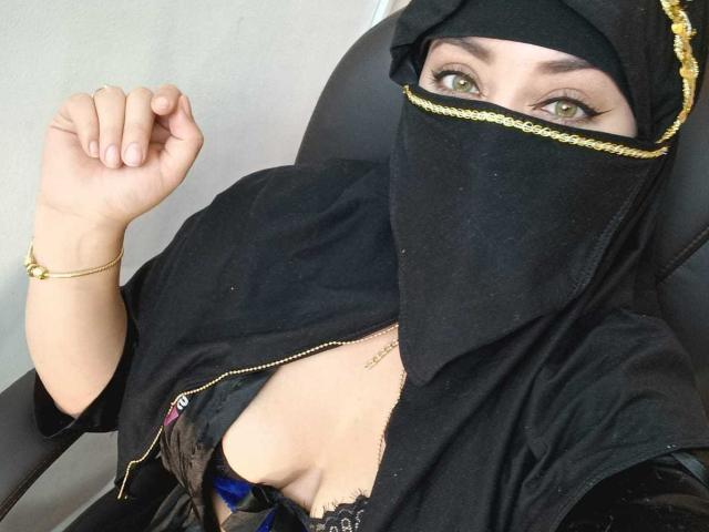 ZahraJalid in black hijab with gold trim, black mask, showing green eyes, gold bracelet, and black lace bra, raised fist, seated in chair, exposing her tits.