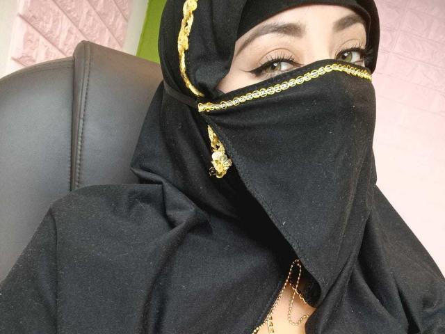 ZahraJalid with black hijab, gold chain, and black mask covering her nose and mouth, showing green eyes and a small glimpse of her cleavage. Sitting on a black leather chair.