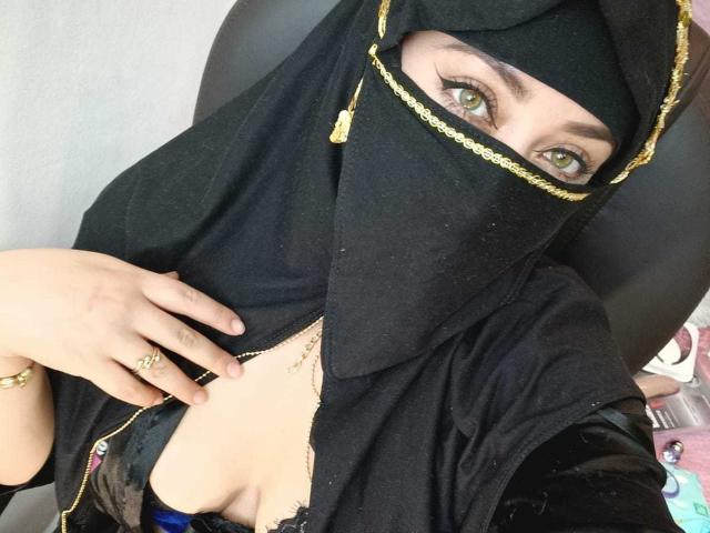 ZahraJalid wearing a black hijab with gold chain, her green eyes peeking out, showing cleavage in a blue lace bra, her hand with rings touching her chest.