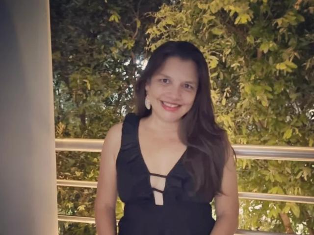 EstelaHot with long brown hair, smiling, wearing a black dress with a deep cleavage and lace-up front, standing outside at night, green leaves in background.
