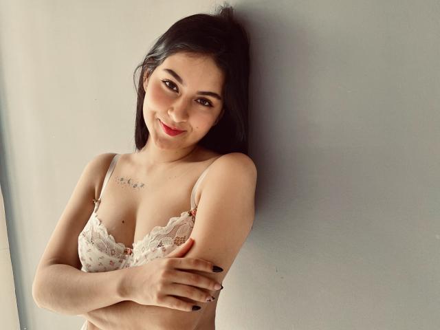 IrinaCarter with dark hair, wearing a white lace bra, arms crossed, smiling at the camera, small star tattoo above her left breast, fair skin, black nail polish.