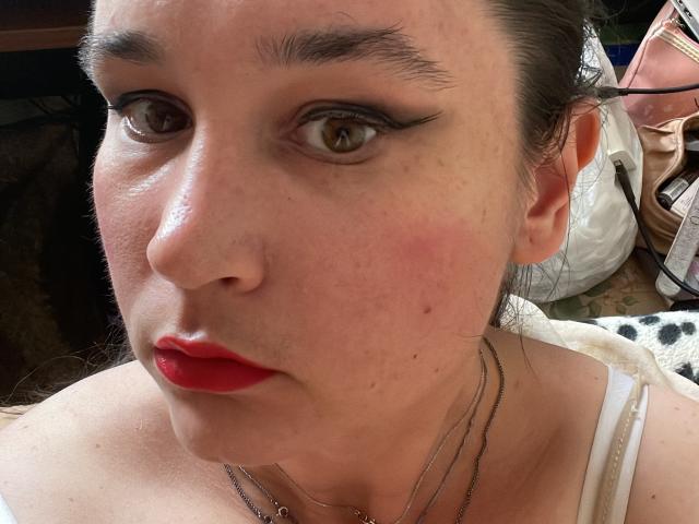 SaliBerry has red lipstick, dark eyeliner, and pale skin with freckles. She's wearing a white top and multiple necklaces, with a pink bag in the blurry background.