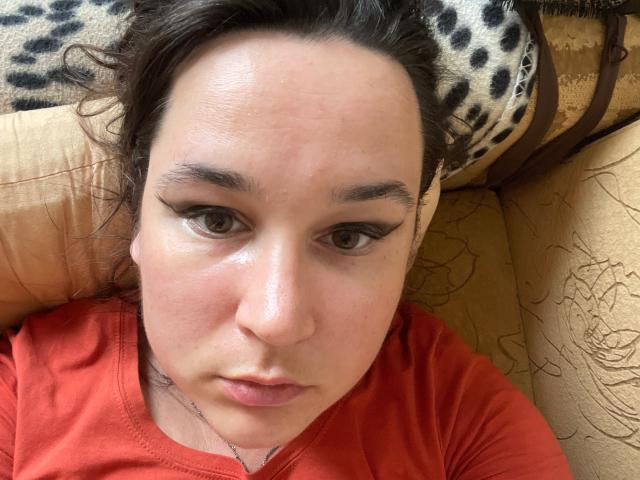 SaliBerry has a serious face, brown eyes with thick black eyeliner, and is lying on a couch in a red shirt. She has a small nose and slightly parted lips.