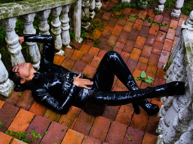 SteffyMiller is lying on a brick path, wearing a shiny black latex bodysuit and high heels, with hands on her belly, legs spread, and eyes closed.
