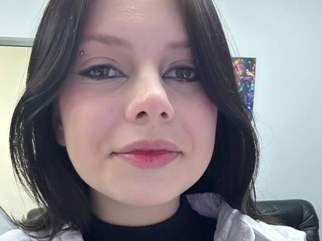 O rosto de RenataSweety, com cabelo preto, pele clara, uma tatuagem discreta de sobrancelha preta e lábios rosados, vestindo uma camisa branca por cima de um gola alta preta, olhando para a câmera..