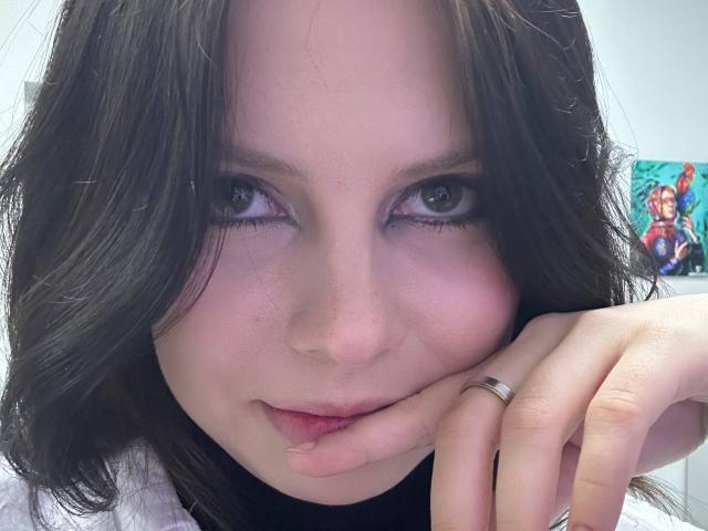 RenataSweety, avec ses cheveux bruns foncés ondulés, sa peau claire et ses yeux bleus, appuyant son menton sur sa main, portant une bague en argent et un pull noir à col roulé..