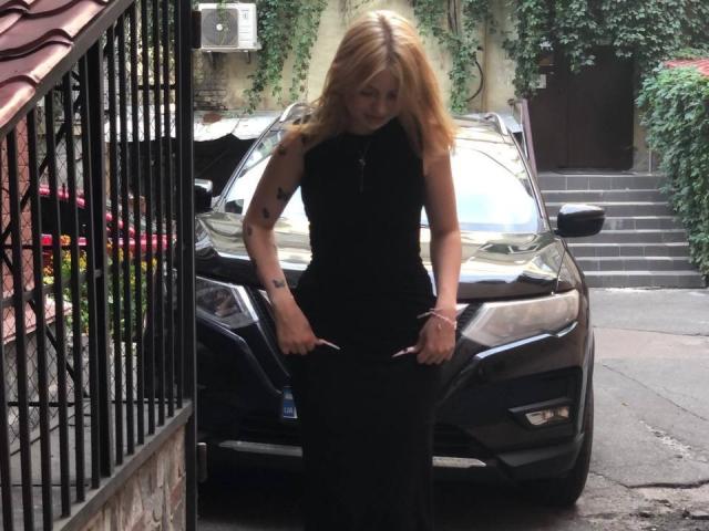 RenataSweety with blonde hair, black sleeveless dress, standing in front of a black car, hands on her hips, visible tattoos on her arms, outside in a narrow alley.