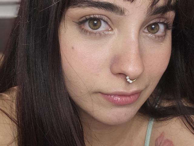 AshleyDaphna tiene ojos marrones, un piercing en la nariz con una pequeña joya y cabello lacio y oscuro. Está mirando a la cámara con los labios ligeramente entreabiertos y de color rosa..