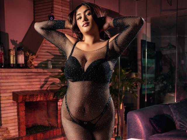 SarhaCooperX in a black mesh bodysuit with sparkling details, showing her big tits and belly, wearing a black bra and thong, standing with hands behind head.