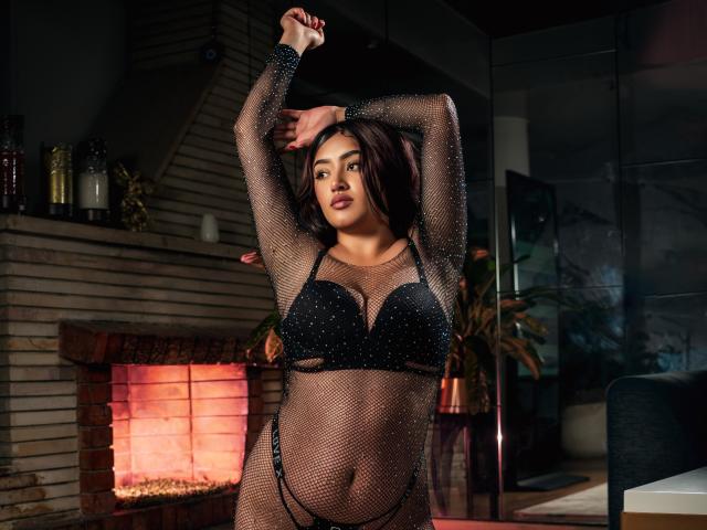 SarhaCooperX with dark brown hair, wearing a black mesh long-sleeve top and black bra, arms raised, showing her tits and small black panties, standing in front of a fireplace.