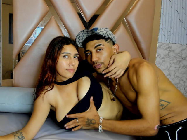 AnyelinAndMark with small tits, in a black top, and Mark, shirtless, with a tattoo on his side, holding her tits, leaning in, both looking at the camera.