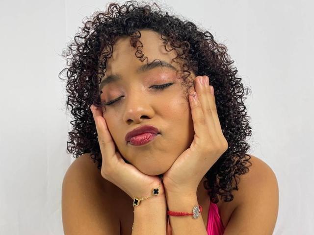 MichelBigTits with curly dark hair, closed pink-shaded eyes, and pink lipstick, hands on cheeks, wearing a pink halter top, red bracelet, and black ring.
