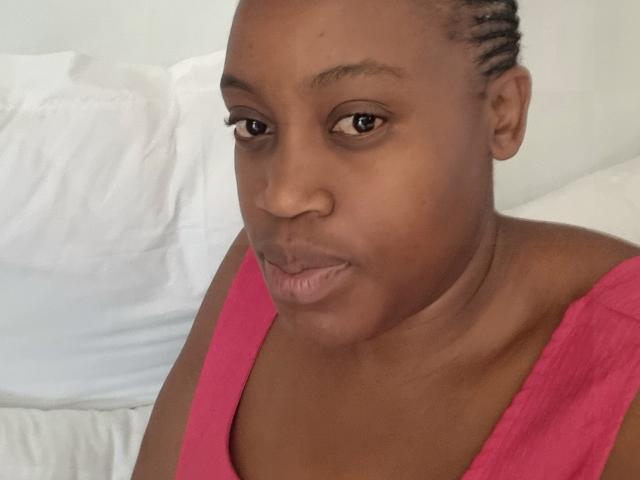 WildBigAss has dark brown skin, short braided hair, and wears a pink top. She's looking at the camera with a neutral expression, showing her small, unadorned lips.
