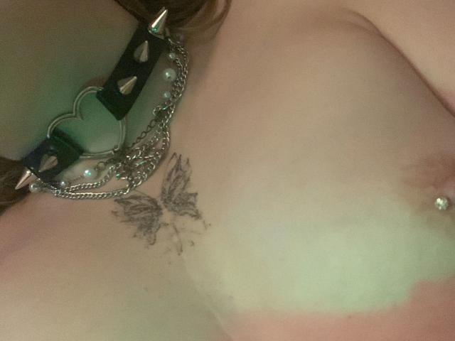 CherryTease-hot's bare tits with a green leather heart-studded collar, silver chain, butterfly tattoo, and nipple piercing in a slightly blurry photo.