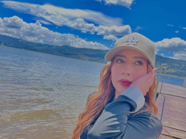 CharlotteAristizabal with long red hair, wearing a gray baseball cap with "LA" and a black jacket, resting her chin on her hand, standing by a wooden dock.