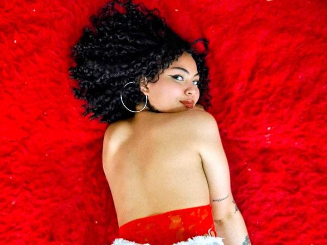 SammyHills69, con cabello negro rizado, mirando por encima del hombro, vestido rojo sin mangas, aros grandes, tatuajes en el brazo derecho, acostada sobre una superficie roja y esponjosa, sin ropa en la espalda, sexy..